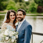 A smiling bride and groom stand together outdoors by a serene lake. The bride holds a bouquet of white and light blue flowers and wears a lace wedding dress with a veil, while the groom is dressed in a blue suit and tie. Green trees are visible in the background, captured beautifully by their Maidstone wedding photographer. Image by Pearce Wedding Photography.
