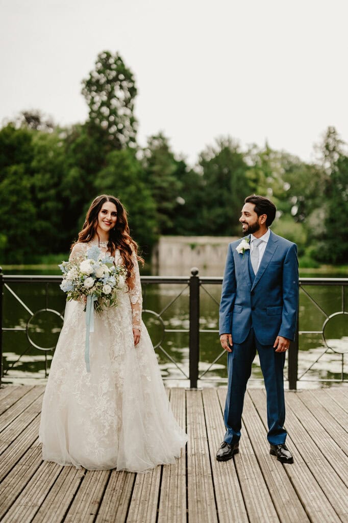 A bride in a white gown holding a bouquet stands beside a groom in a blue suit. They are on a wooden deck with a background of trees and a body of water.
