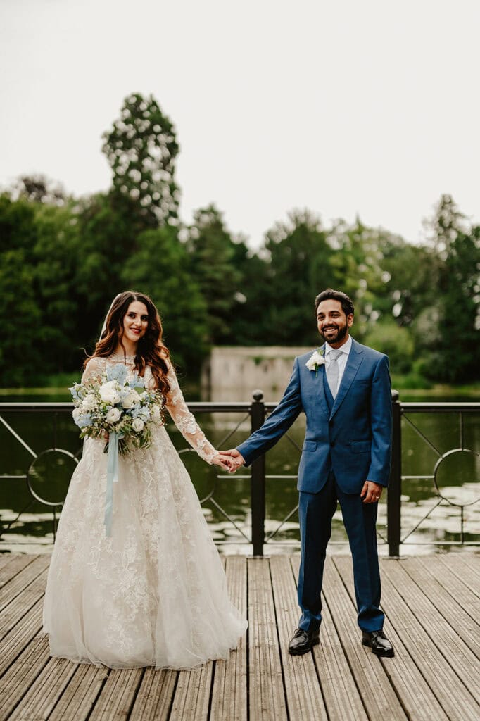 A bride and groom stand on a wooden deck, holding hands and facing the camera. The bride is in a white dress holding a bouquet, and the groom is in a blue suit. Trees and a lake are in the background.