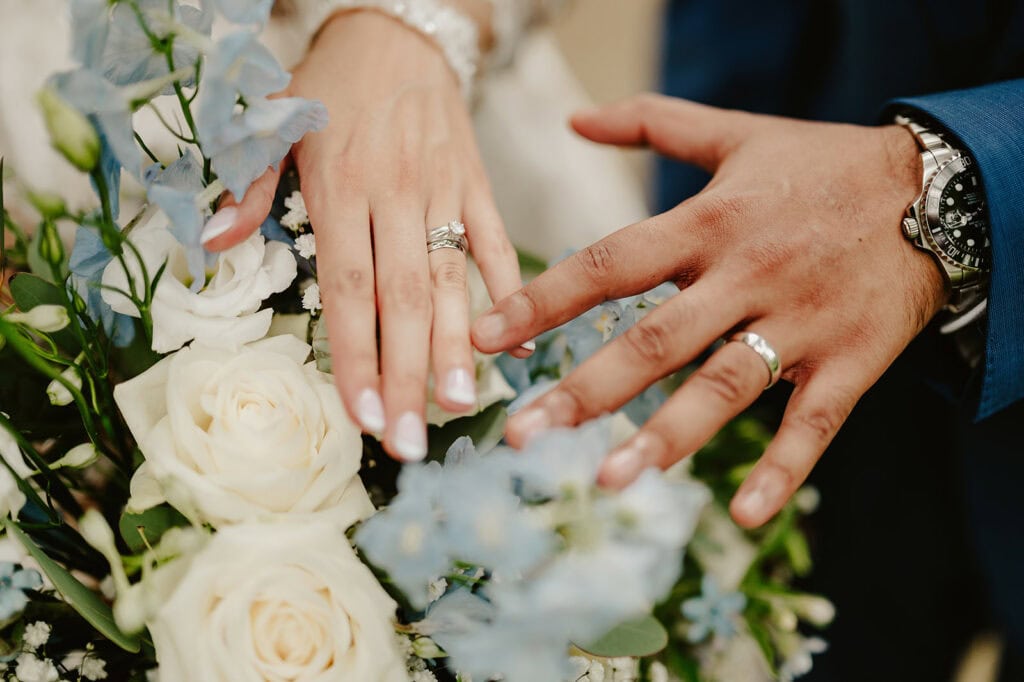 Close-up of a couple's hands showing wedding rings, captured beautifully by a Maidstone wedding photographer. The hands rest above a bouquet featuring white roses and light blue flowers. The bride's nails are manicured, and she is wearing a bracelet, while the groom is adorned with a watch. Image by Pearce Wedding Photography.