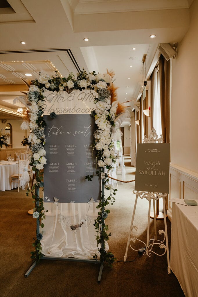A wedding seating chart stand decorated with white flowers and greenery, positioned next to a sign with gold text on an easel in a well-lit, elegantly decorated room with round tables.