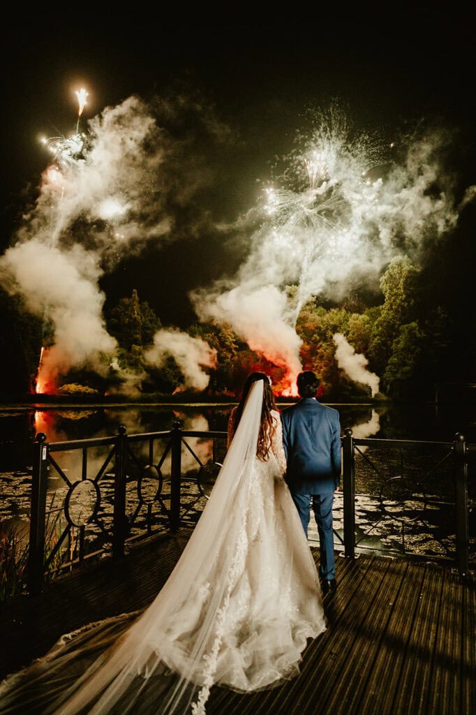 A bride and groom stand on a dock at night, watching a fireworks display over a lake surrounded by trees. The bride wears a long veil; the groom is in a light blue suit.