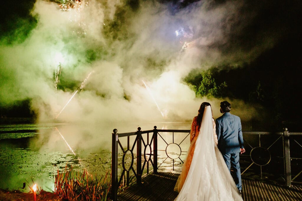 A bride and groom stand on a wooden deck watching a firework display over a misty lake at night.