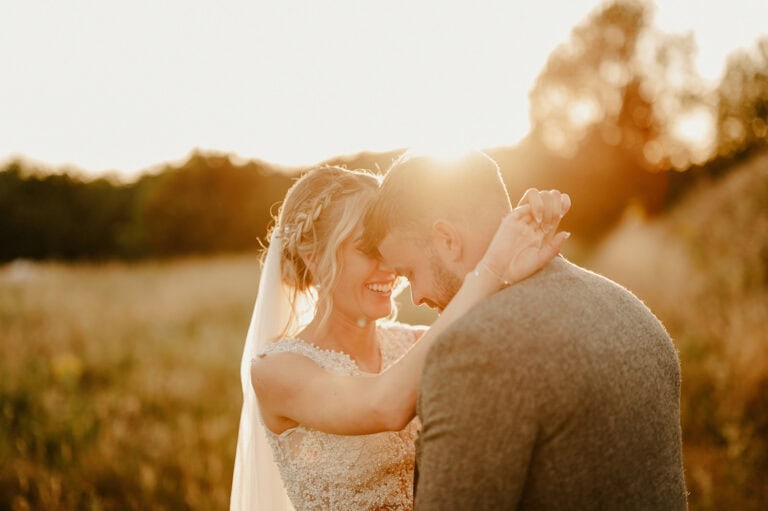 A bride and groom embrace in a sunlit field near Villiers Barn during golden hour. The bride, in a lace gown and veil, smiles while holding the groom's head. The groom, in a gray suit, leans in softly. Sunlight glows warmly behind them, casting a serene glow over the picturesque scene. Image by Pearce Wedding Photography.