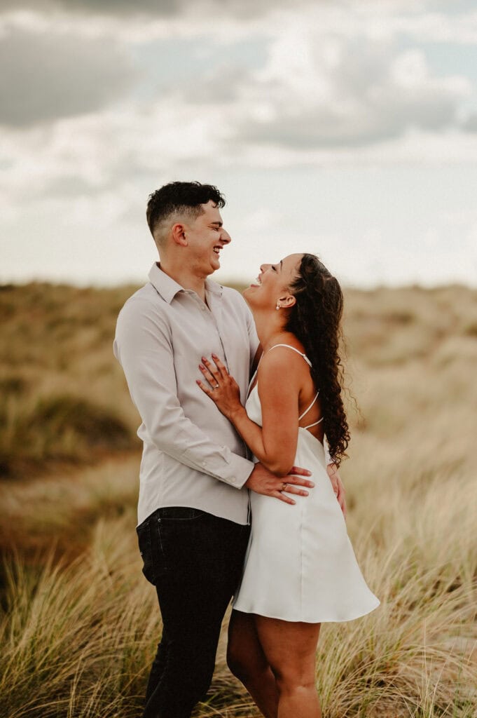 A couple stands closely together in a field, both smiling and gazing at each other. The woman wears a white dress, and the man wears a light-colored shirt and dark pants.