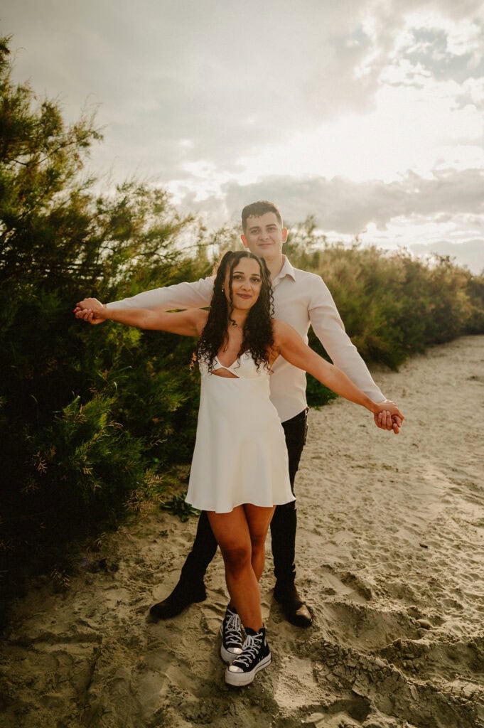 A couple stands on a sandy path surrounded by greenery, capturing a perfect pre-wedding moment. The woman, in a white dress and black sneakers, holds hands with the man behind her in a white shirt. They both smile under a partly cloudy sky. Image by Pearce Wedding Photography.