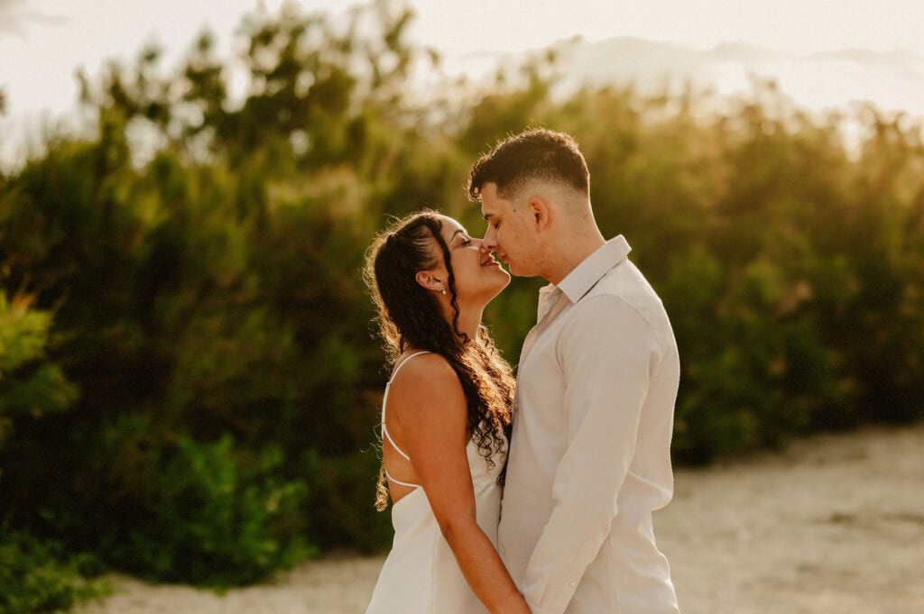A couple, both dressed in light clothing, stand close together and share a kiss in an outdoor setting with greenery in the background.