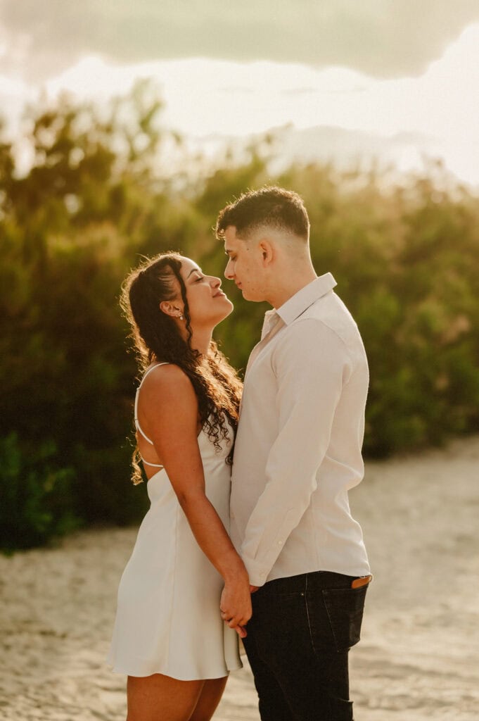 A couple stands closely together on a sandy beach, holding hands and looking at each other, with greenery in the background and sunlight filtering through the trees.