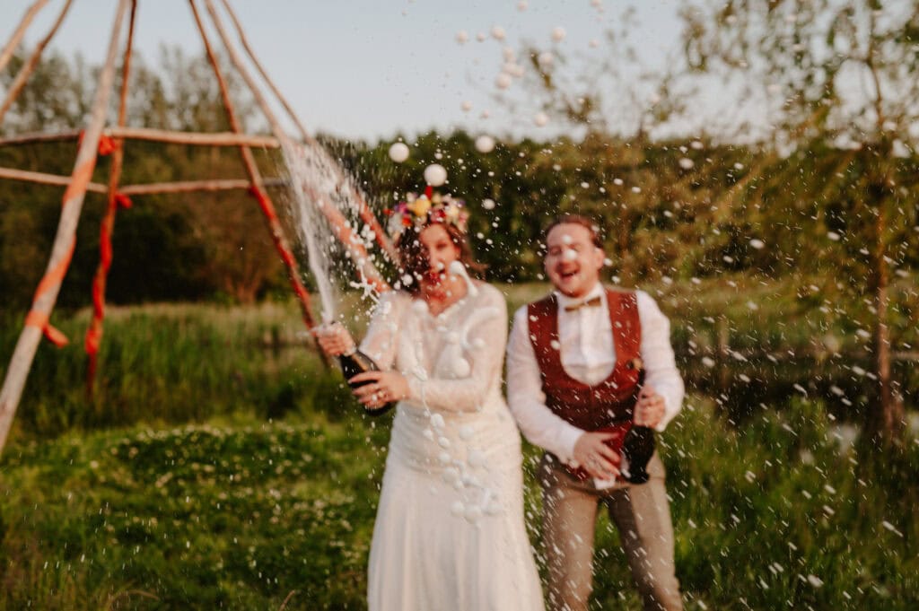 A bride and groom in wedding attire pop champagne bottles outdoors, with champagne spraying out energetically. They are both smiling and appear to be celebrating. Trees and a structure are visible behind them.