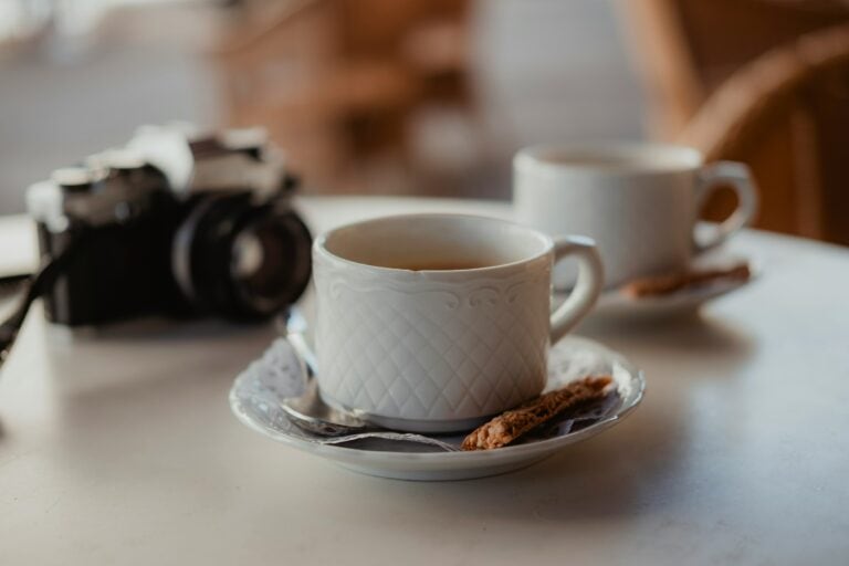 Two white ceramic cups with saucers and spoons on a table, accompanied by small cookies and a vintage camera in the background.