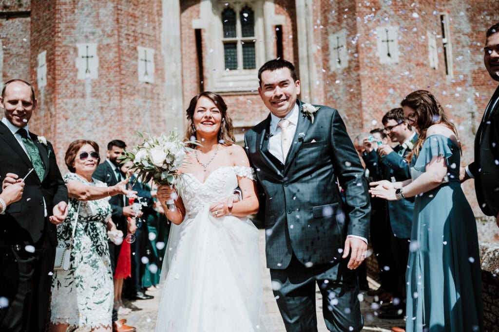A bride and groom, smiling, walk down a path outside the historic Herstmonceux Castle as guests throw confetti. The bride holds a bouquet, and the groom is in a grey suit, making their wedding day unforgettable.