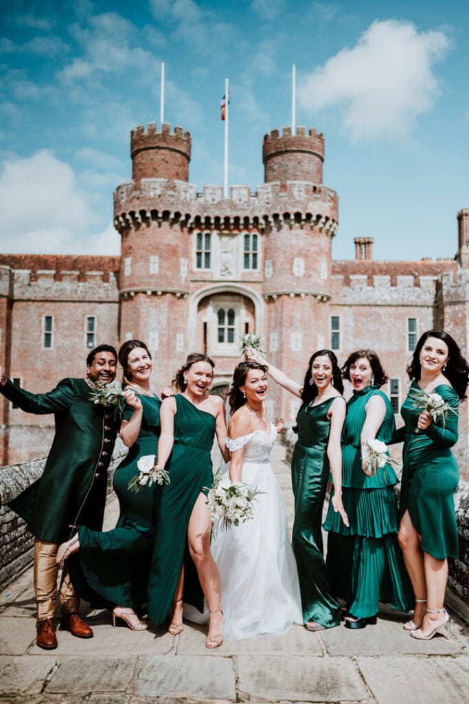 A bride in a white dress stands with bridesmaids and a groomsman in green outfits, posing cheerfully in front of the historic Herstmonceux Castle, creating an enchanting wedding scene.