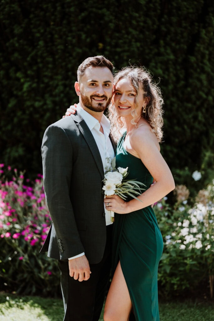 A man in a dark suit and a woman in a green dress with a leg slit stand closely together, smiling, in front of the lush garden at Herstmonceux Castle.