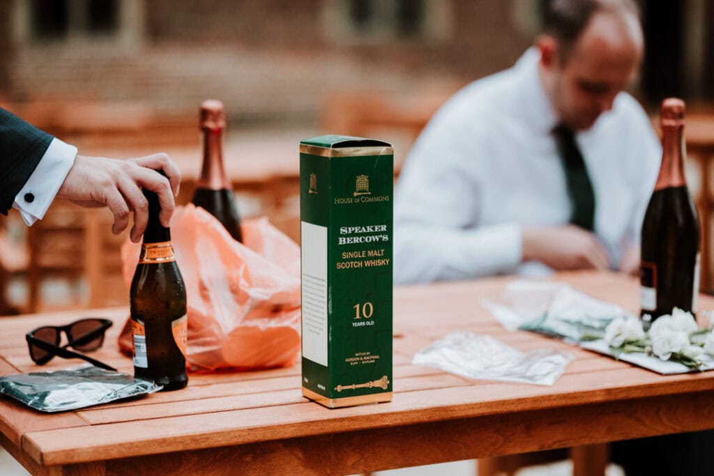 A table with a green box labeled "Speaker Bercow's Single Malt Scotch Whisky," a beer bottle, sunglasses, and other items. At Herstmonceux Castle, two people are seated at the table during a wedding, with one reaching for the beer bottle.