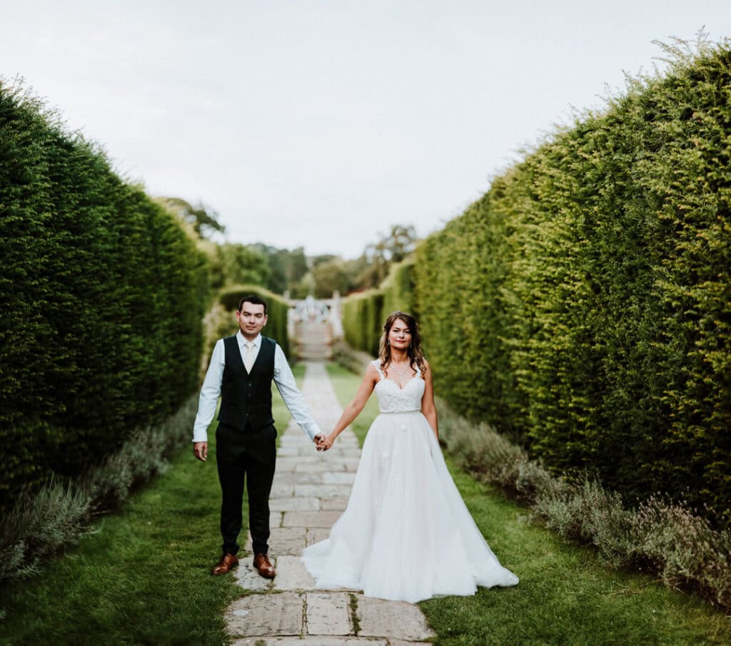 A couple holding hands stands on a stone pathway surrounded by tall green hedges, reminiscent of the enchanting gardens of Herstmonceux Castle. The woman is wearing a flowing white dress, and the man is in a crisp black vest and trousers with a white shirt, perfect for their wedding day.