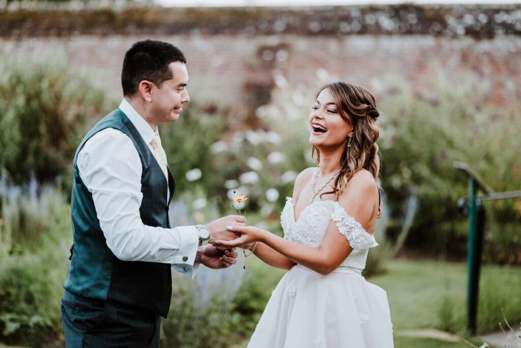 A bride and groom smile and interact with each other while standing in a garden at Herstmonceux Castle. The bride wears a white dress, and the groom wears a vest and tie, creating a picturesque wedding scene.