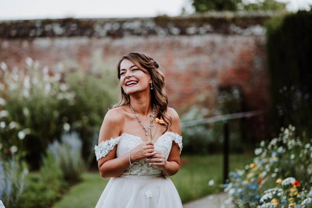 A smiling woman in an off-shoulder white dress stands outdoors in front of a brick wall, holding a flower, reminiscent of a serene moment at Herstmonceux Castle on her wedding day.