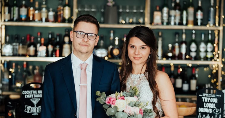 A couple stands in front of a bar, shelves stocked with liquor bottles. The man wears a dark suit with a light pink tie, and the woman is in a light-colored dress holding pink and white flowers, perfectly capturing the romance of their special day at the Albion House, renowned as a premier Thanet wedding venue. Image by Pearce Wedding Photography.