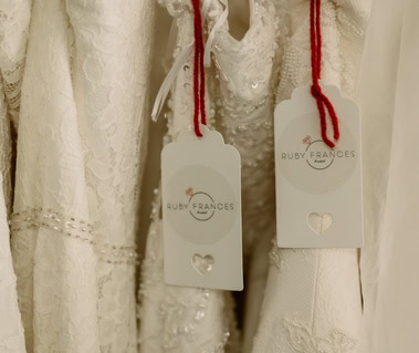 A close-up of two white lace garments with red stringed tags labeled "Ruby Frances" hanging side by side in a quaint Thanet bridal shop.