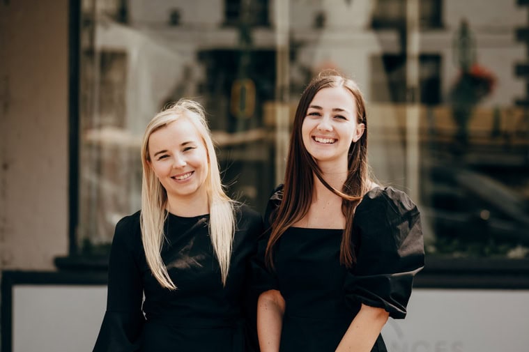 Two women with long hair, both dressed in black tops, are standing side by side smiling in front of a window of a bridal shop in Thanet.