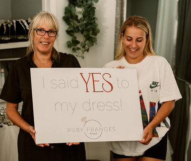 Two women hold a sign that says, "I said YES to my dress!" with the Ruby Frances bridal shop logo at the bottom. Both are smiling; one wears glasses and a dark top, the other is in a white T-shirt with a Nike logo, celebrating their special moment in Thanet.