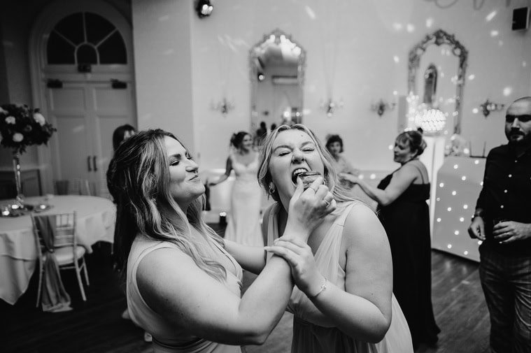 Two women are playfully interacting at a wedding celebration, with one woman feeding the other a piece of food. A relaxed atmosphere fills the decorated event hall, while other attendees and the photographer are seen in the background capturing the joyous moments.