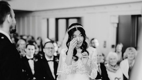 A bride wipes a tear from her eye during a relaxed wedding ceremony, while guests watch and the groom stands nearby. The photographer captures this tender moment in black and white.