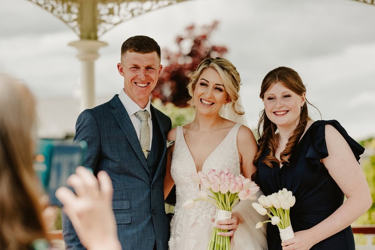 Three people are posing for a relaxed photo at an outdoor event. One person is dressed in a dark suit, one in a white dress holding pink flowers, and one in a dark dress holding white flowers, capturing the essence of the wedding beautifully for the photographer.