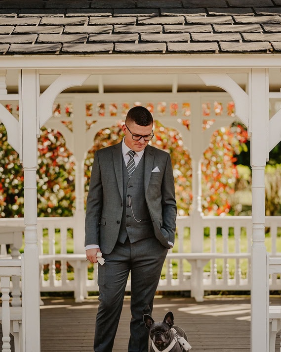 A relaxed man in a grey suit stands under a white gazebo at a wedding, holding a leash attached to a small dog.