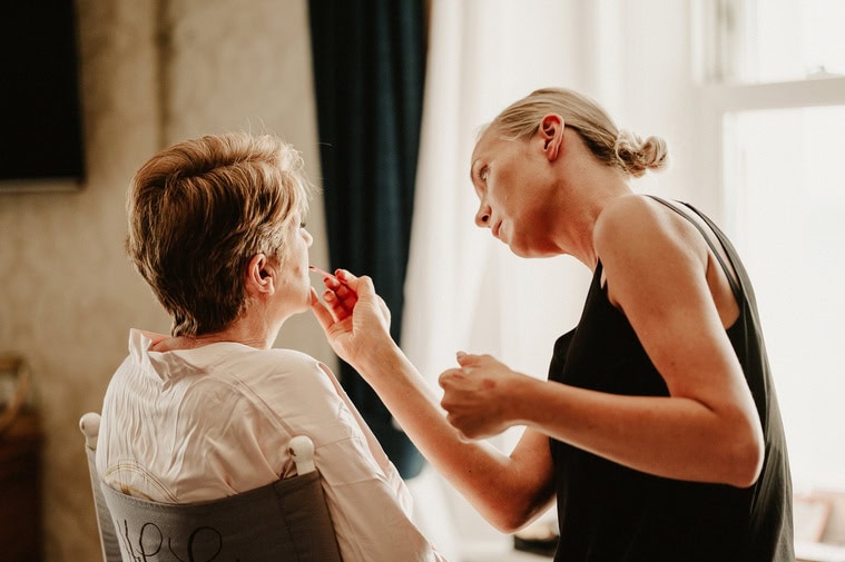 In a well-lit room with a curtain and window in the background, a woman is applying makeup to another woman seated in a chair, looking relaxed as they prepare for the wedding. Off to the side, a photographer captures the serene moment.