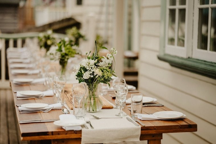 A long wooden table set for a formal meal, adorned with white plates, glassware, cutlery, light-colored napkins, and flower arrangements crafted by a local florist in glass jars, is on an outdoor patio next to a building in Kent.
