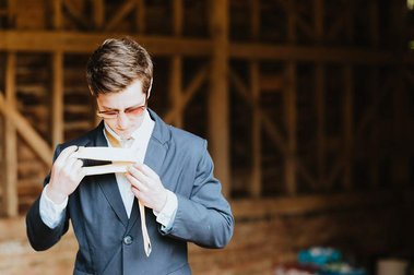 A man in a suit and glasses stands in a wooden structure, reminiscent of an old papermill, holding and inspecting a yellow necktie, perhaps preparing for a wedding.