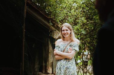 A woman in a floral dress stands outside near the greenery of a Kent garden, smiling with her arms crossed, as if reminiscing about a lovely wedding moment.