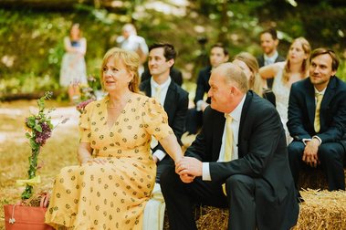 A couple sits holding hands on hay bales during a charming outdoor wedding. The woman, in a yellow floral dress, and the man, in a suit with a matching tie, smile warmly at each other. In the background, guests near the historic Kent papermill look on with joy.
