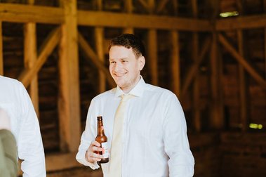 A man in a white shirt and tie holds a beer bottle, standing in a wooden structure with beams, perhaps at a wedding venue in Kent.