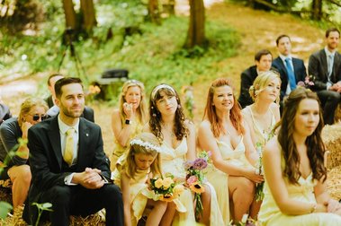 A group of people, including women in yellow dresses and a man in a suit, are seated outdoors on straw bales during a daytime wedding at Papermill Creek in Kent's wooded area.