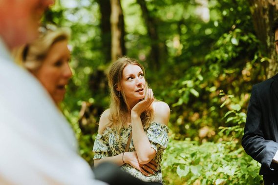 A woman in a floral dress stands outdoors in a wooded area at a Kent wedding, resting her chin on her hand and looking thoughtfully to the side. Other people are partially visible around her.