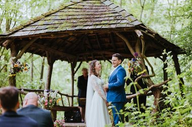 A couple stands under a rustic wooden gazebo in a forest, exchanging vows during their wedding ceremony. Guests are seated, watching the event while flowers decorate the structure, lending an air of natural elegance one might find in Kent or near an old papermill.
