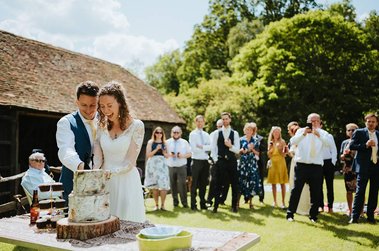 A couple in wedding attire cuts a cake outdoors on a sunny day in Kent, surrounded by guests who are watching and taking photos.