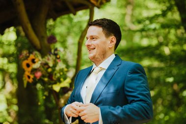 A man in a blue suit stands outside, smiling, with greenery and blurred flowers in the background, as if captured at a lush wedding venue in Kent.