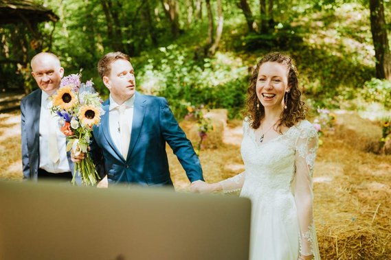 A man in a navy suit and a woman in a white wedding dress hold hands outdoors, with another man in a suit standing next to them. They are all smiling, surrounded by trees and hay bales with flowers—a perfect Kent setting for their special day.