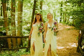 Two women in yellow dresses walk down a wooded path holding flower bouquets, surrounded by lush greenery, as they head towards the charming Kent papermill for a wedding celebration.