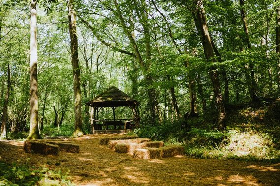 A wooden gazebo stands amidst a forest with sunlight filtering through the trees in Kent. Hay bales are arranged in rows in front of the gazebo, creating a rustic seating area perfect for a wedding.