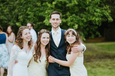 A man and three women, all dressed in formal attire, stand outdoors posing for a photo at a wedding. There is greenery in the background, and other people are visible in the distance, enjoying the beautiful day in Kent.