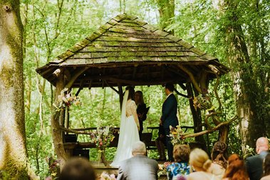 A couple stands at an altar under a rustic wooden gazebo in a serene Kent forest setting, exchanging vows during an enchanting outdoor wedding ceremony, while guests are seated watching.