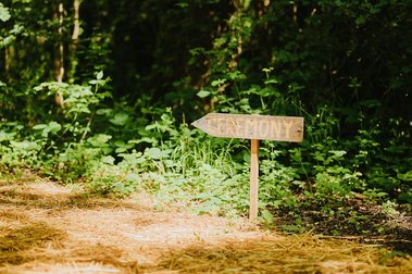 A wooden sign labeled "CEREMONY" points to the right, situated in a picturesque forested area in Kent. Sunlight filters through the trees, highlighting the serene path to a beautiful wedding celebration ahead.