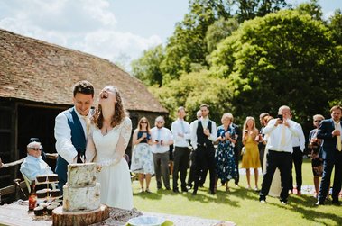 A bride and groom smile while cutting a wedding cake outdoors at the Papermill in Kent on a sunny day, surrounded by guests watching and taking photos.