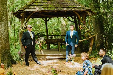 Two men in suits stand in front of a rustic wooden gazebo in a forest, one holding a floral bouquet. Seated guests in summer attire enjoy the wedding scene, adding to the charming atmosphere that feels like it's straight out of Kent.