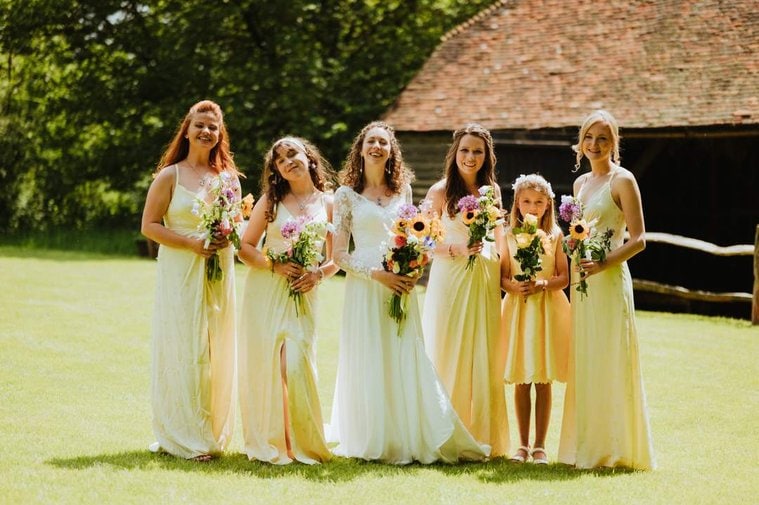 A bride stands outside with five bridesmaids, all wearing light yellow dresses and holding flower bouquets, posing in front of a rustic building at a charming Kent wedding.