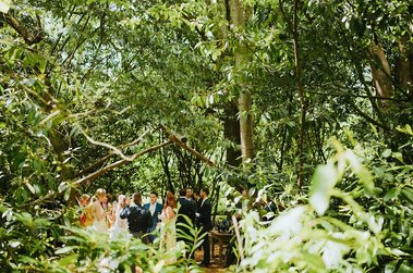 A group of people is gathered for a wedding ceremony in a forest in Kent. The bride and groom stand under a wooden archway surrounded by trees and greenery, creating a magical atmosphere for their special day.
